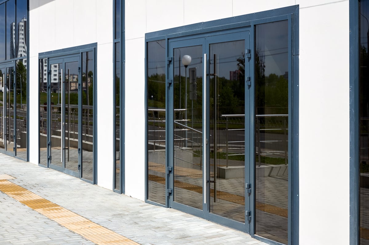 Dark gray aluminum double doors with large glass panels installed in white building facade. Modern architectural design features long vertical handles and matching side windows along paved sidewalk.